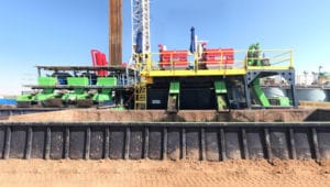 A large industrial drilling rig with green, red, and yellow components, set on a sandy construction site under a clear blue sky.